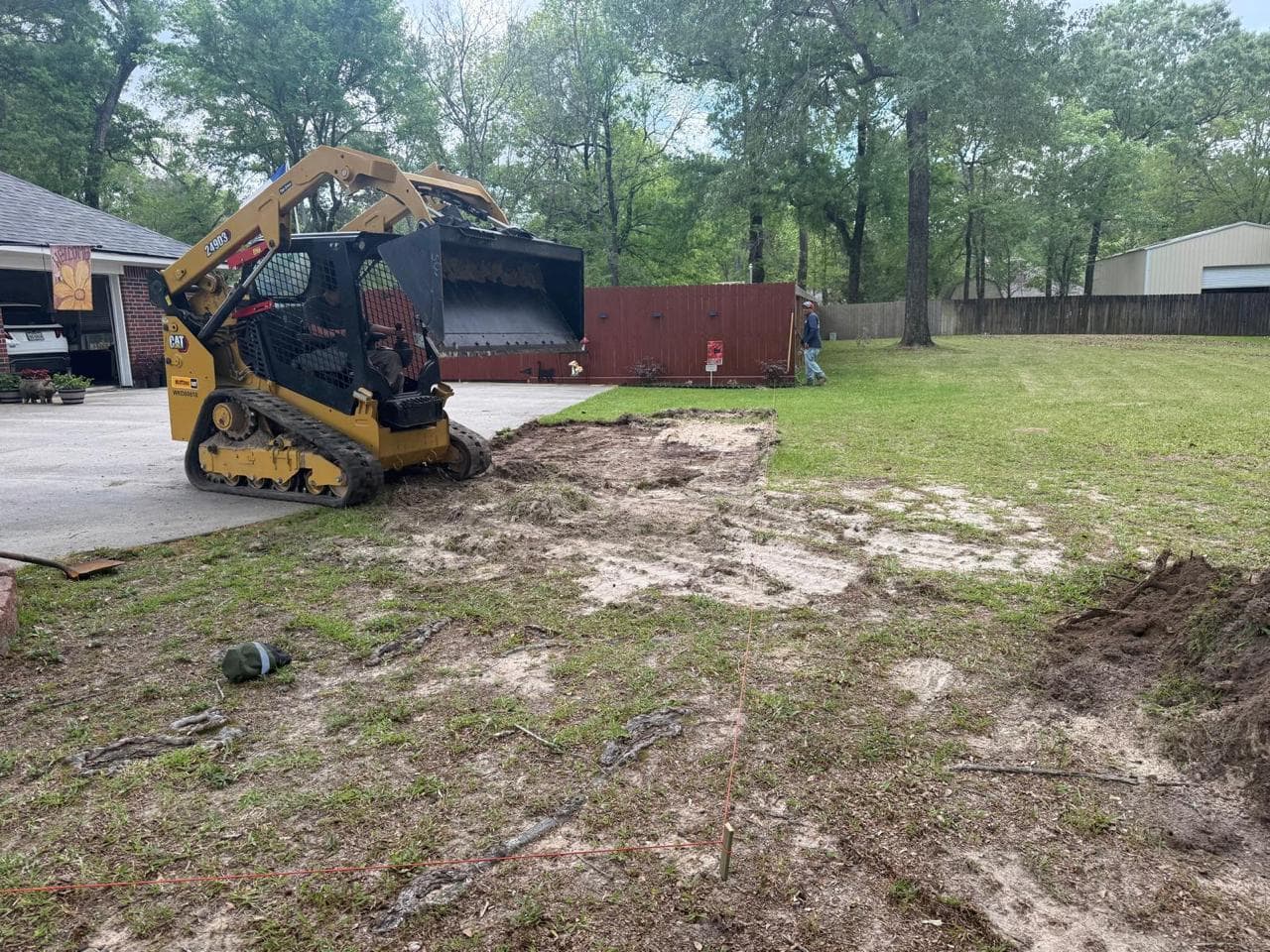 Tracked skid steer clearing surface vegetation and grading alongside a driveway